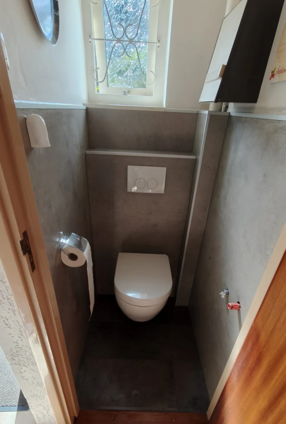 Hetzelfde toilet na renovatie: hangtoilet met voorzetwand en grijze betonlook wandpanelen onder hetzelfde raampje