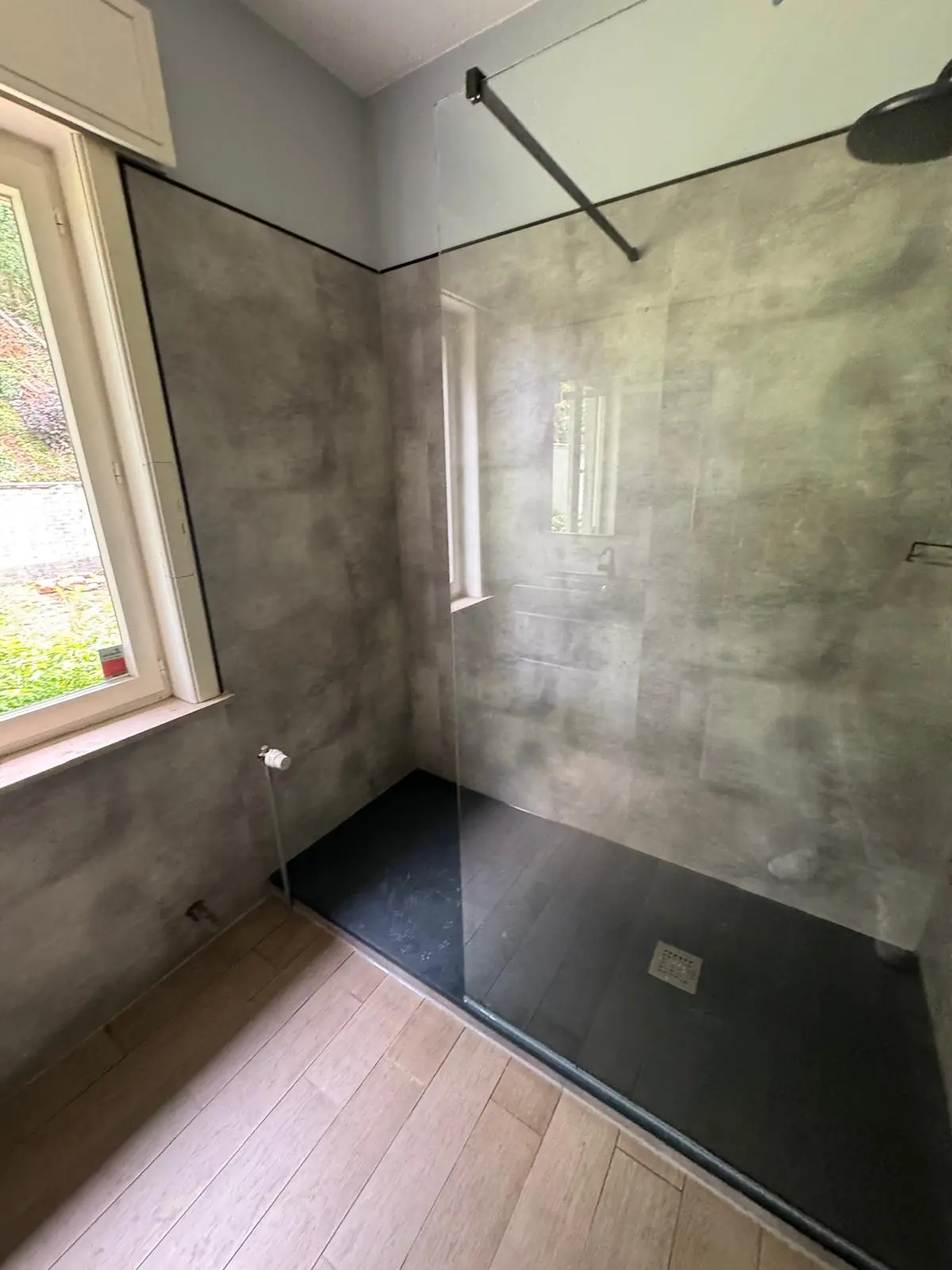 Inloopdouche met grijze betonlook wandpanelen, zwarte douchebak en glazen wand naast raam