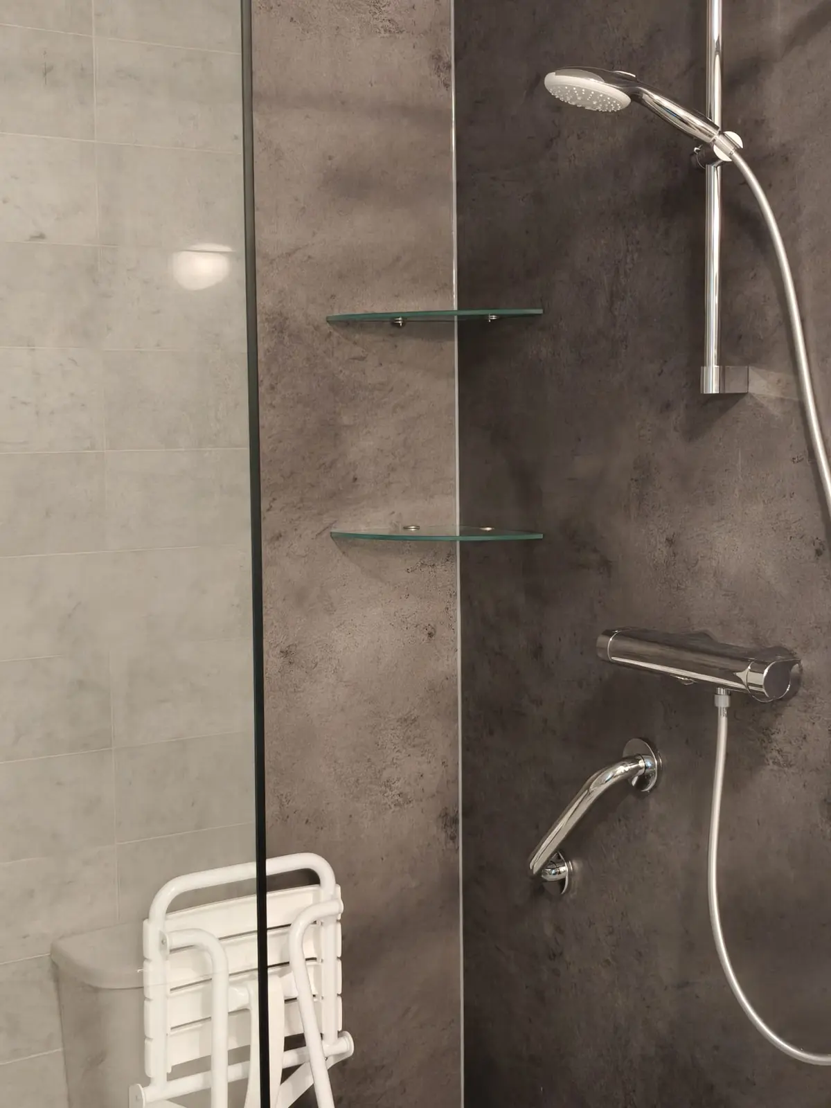 Douche met donkere betonlook wandpanelen, glazen planchets, wandbeugel, thermostaatkraan en douchezitje